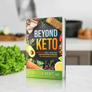 Dr. Colbert’s Beyond Keto Hardcover book by Dr. Don Colbert, MD from Keto Zone displayed standing on a kitchen countertop with fresh vegetables and healthy foods in the background. The book focuses on a Mediterranean-Keto lifestyle designed to support fat burning, gut health, metabolic balance, and digestion through nutrient-dense foods, probiotics, fiber-rich nutrition, and healthy fats such as MCT oil.