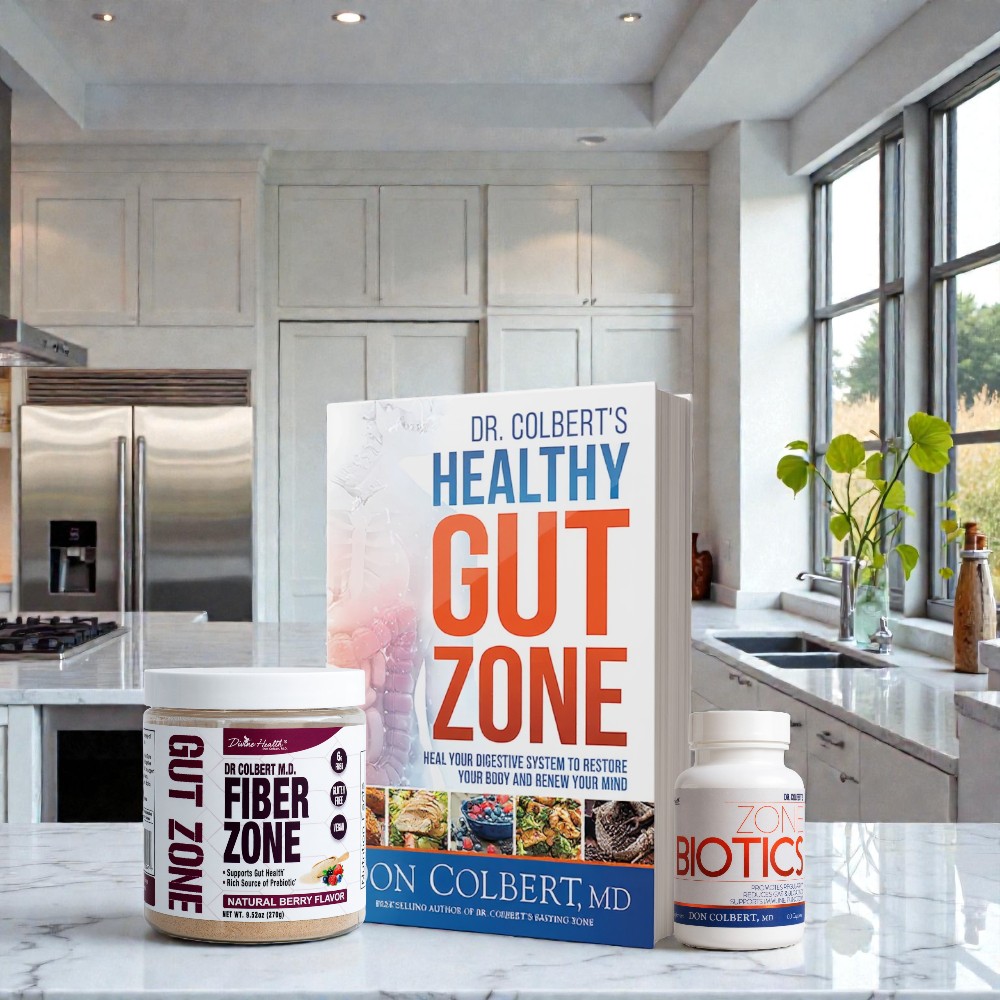 Divine Health Dr. Don Colbert, MD Gut Zone Starter Pack arranged on a polished marble kitchen countertop, featuring a Fiber Zone container in Natural Berry Flavor with maroon-and-white labeling, Dr. Colbert’s “Healthy Gut Zone” book displayed upright in the center with bold blue and orange title text, and a white bottle of Zone Biotics positioned to the right. The background includes a modern kitchen setting with stainless-steel appliances, white cabinetry, large windows, natural greenery, and bright daylight.
