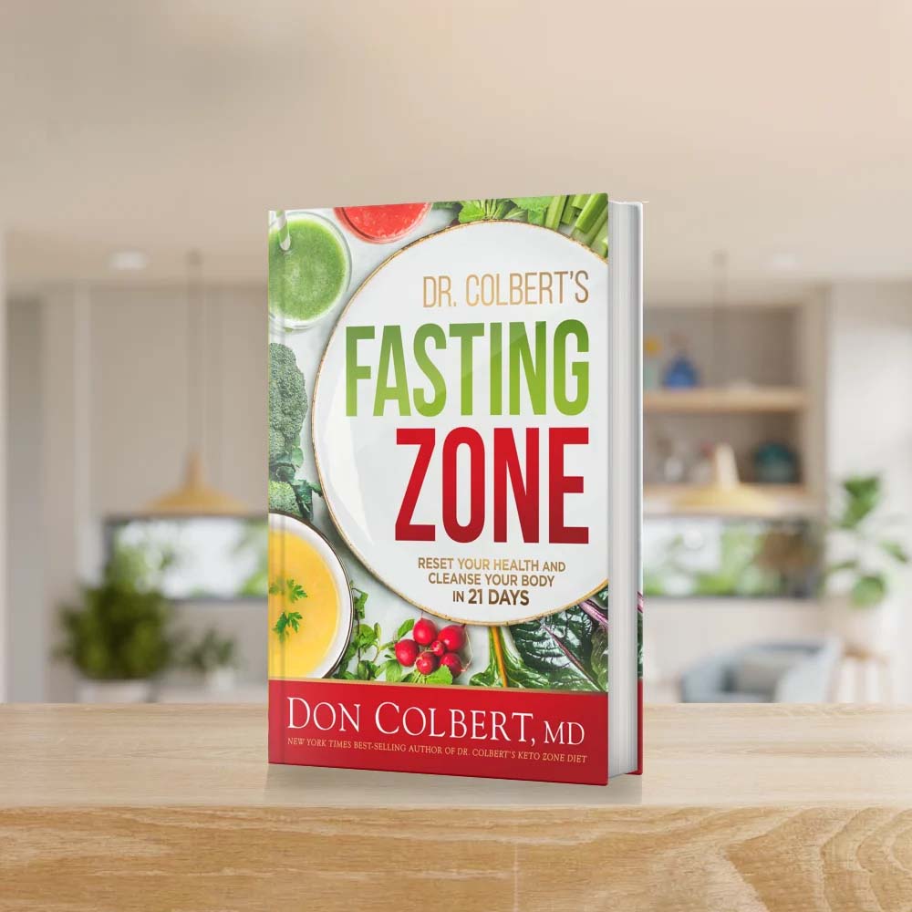 Dr. Colbert’s Fasting Zone book by Dr. Don Colbert, MD from Keto Zone displayed standing on a wooden kitchen counter with fresh vegetables and green foods in the background. The book presents a 21-day fasting and nutrition program designed to support gut health, metabolism, detoxification, and digestive balance while emphasizing probiotics, fiber-rich foods, healthy fats like MCT oil, and nutrient-dense nutrition for overall wellness.