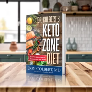 A hardcover copy of Dr. Colbert’s Keto Zone Diet standing upright on a polished wooden kitchen countertop. The cover features a rustic food-themed design showing salmon, lemon slices, herbs, peppers, and spices arranged on a wooden surface. The title “Dr. Colbert’s Keto Zone Diet” appears in large bold letters, with a red banner displaying the original tagline. The author’s name, Don Colbert, MD, appears at the bottom along with his New York Times bestselling designation. The background shows a bright, modern kitchen with natural light, blurred décor, and neutral tones, giving the product a clean lifestyle presentation.