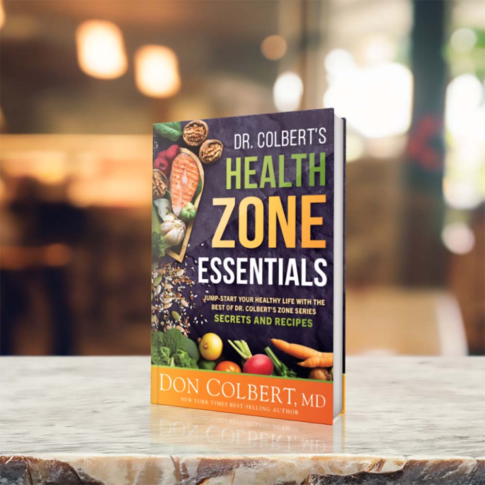Dr. Colbert’s Health Zone Essentials book by Dr. Don Colbert, MD from Divine Health displayed upright on a marble countertop with a softly blurred café-style background. The wellness guide focuses on foundational health strategies including gut health support, balanced nutrition, and lifestyle improvements. It highlights how digestive support through probiotics, fiber supplements, collagen-supporting foods, and nutrition strategies that may include MCT oil powder can help support overall wellness and metabolic balance.