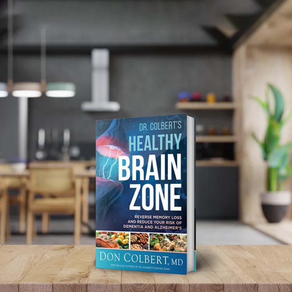 Dr. Colbert’s Healthy Brain Zone book by Dr. Don Colbert, MD from Divine Health displayed upright on a wooden table in a modern kitchen setting. The book focuses on supporting brain health, memory, and cognitive function while highlighting the connection between gut health, probiotics, fiber-rich nutrition, and healthy fats such as MCT oil. The guide provides strategies for supporting mental clarity and long-term neurological wellness through nutrition and lifestyle choices.