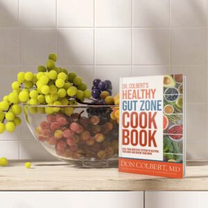 Dr. Colbert’s Healthy Gut Zone Cookbook by Dr. Don Colbert, MD from Divine Health displayed on a kitchen countertop beside a glass bowl of fresh grapes. The cookbook focuses on gut health nutrition and recipes designed to support digestive balance and microbiome health. It highlights foods that naturally support probiotics, fiber intake, and collagen-supporting nutrition while complementing wellness approaches that may include fiber supplements, probiotics, and MCT oil powder for improved digestive health and overall vitality.