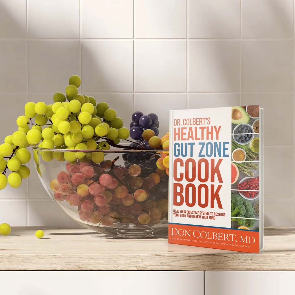 Dr. Colbert’s Healthy Gut Zone Cookbook by Dr. Don Colbert, MD from Divine Health displayed on a kitchen countertop beside a glass bowl of fresh grapes. The cookbook focuses on gut health nutrition and recipes designed to support digestive balance and microbiome health. It highlights foods that naturally support probiotics, fiber intake, and collagen-supporting nutrition while complementing wellness approaches that may include fiber supplements, probiotics, and MCT oil powder for improved digestive health and overall vitality.