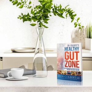 Dr. Colbert’s Healthy Gut Zone hardcover book by Dr. Don Colbert, MD from Divine Health displayed on a modern kitchen counter beside a coffee cup and decorative plant. The book focuses on gut health and digestive wellness, explaining how probiotics, fiber supplements, microbiome balance, and nutrition strategies may help support digestion, immune health, nutrient absorption, and overall wellness through a healthier gut environment.