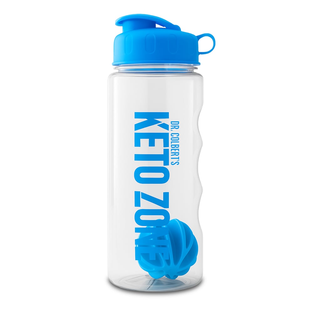 A clear plastic Keto Zone shaker bottle by Dr. Colbert is shown standing upright against a plain white background. The bottle features a bright blue screw-top lid with a flip-up drinking spout and a small loop handle on the side of the cap. Inside the transparent bottle, a matching blue wire shaker ball is visible at the bottom. The front of the bottle displays vertical blue text reading “Keto Zone” with “Dr. Colbert’s” printed alongside it. The bottle has a contoured grip design along one side, giving it a textured appearance. The image is evenly lit, isolated, and focused solely on the bottle with no additional props or background elements.