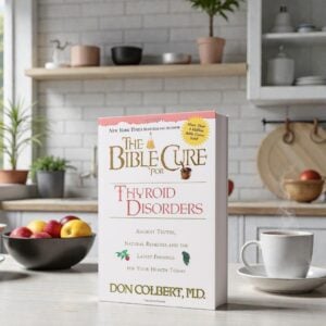 A softcover paperback copy of “The Bible Cure for Thyroid Disorders” by Don Colbert, MD is displayed standing upright on a light-colored kitchen countertop. The book is centered in the frame with its spine visible, showing the thickness of the softcover format. Behind it is a bright, modern kitchen setting with white subway tile backsplash, wooden shelves holding bowls and plants, a cutting board, and neutral décor. A bowl of fresh fruit sits to the left, while a white coffee cup with visible steam rests to the right, creating a warm, natural lifestyle scene.