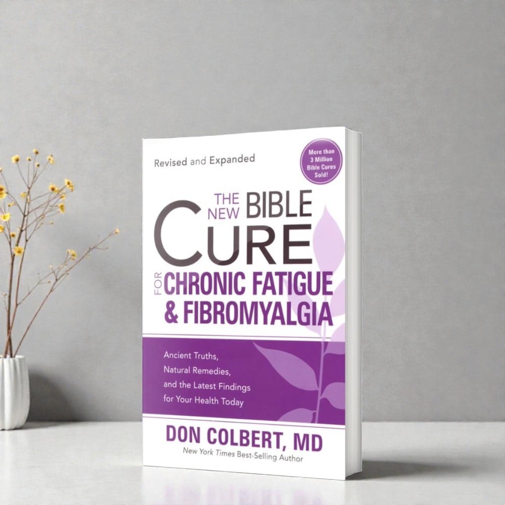 A revised and expanded softcover book titled “The New Bible Cure for Chronic Fatigue & Fibromyalgia” by Don Colbert, MD stands upright on a smooth white tabletop against a light gray wall. The book is angled slightly to the right, showing its softcover binding and page depth. The cover features a purple and white color scheme with a stylized leaf graphic and a circular badge stating “More than 3 Million Bible Cures Sold.” A minimalist ceramic vase holding thin branches with small yellow flowers sits to the left, creating a calm, wellness-focused setting with soft, natural lighting.