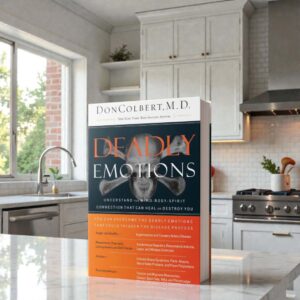 Deadly Emotions book by Dr. Don Colbert, MD from Divine Health displayed standing on a modern kitchen countertop. The book focuses on the connection between emotional health, physical wellness, and spiritual balance, explaining how stress and negative emotions can impact gut health, digestion, immune function, and overall well-being while promoting healthier lifestyle and nutrition habits for long-term wellness.